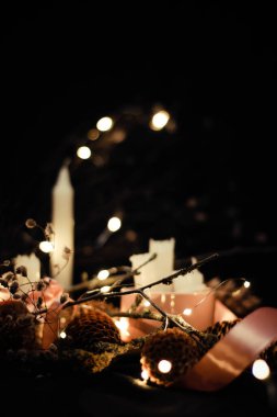 Mum ışığında pembe Noel süslemeleri, pembe kurdeleler, dallar, kuru çimenler, parlak mücevherler, kurdeleler ve çam kozalakları, çiçek fenerleri, bokeh etkisi, siyah bir arka planda.