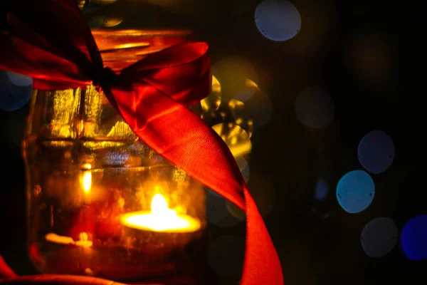 A small candle burns in a jar in the dark against the background of bokeh lights red ribbon bow, bokeh, blurred background, place for text