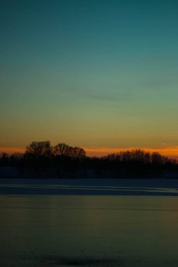 Donmuş gölde gün batımı. Sazlıklar ve ağaçlar, kıyılarda kar.