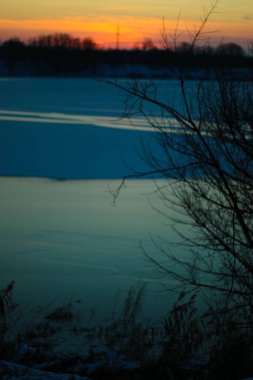 Donmuş gölde gün batımı. Sazlıklar ve ağaçlar, kıyılarda kar.