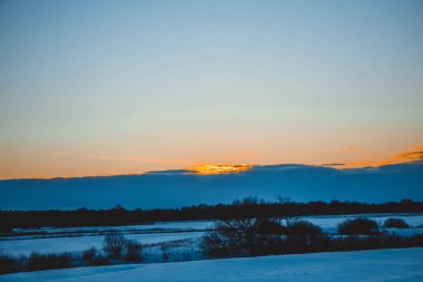 Güzel kış manzarası, bulutlu gün batımı, uzaktaki orman, kardaki tarla ve ağaçlar, dondurucu güneşli bir akşam, Ukrayna