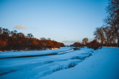 Gün batımında nehirde kış ormanı. Karlı ağaçlarla dolu renkli bir manzara, suda yansıması olan güzel donmuş bir nehir. Mevsimlik. Kış ağaçları, göl ve mavi gökyüzü. Buz gibi karlı nehir. Hava Durumu