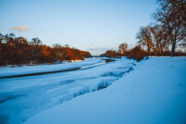 Gün batımında nehirde kış ormanı. Karlı ağaçlarla dolu renkli bir manzara, suda yansıması olan güzel donmuş bir nehir. Mevsimlik. Kış ağaçları, göl ve mavi gökyüzü. Buz gibi karlı nehir. Hava Durumu