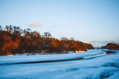 Gün batımında nehirde kış ormanı. Karlı ağaçlarla dolu renkli bir manzara, suda yansıması olan güzel donmuş bir nehir. Mevsimlik. Kış ağaçları, göl ve mavi gökyüzü. Buz gibi karlı nehir. Hava Durumu