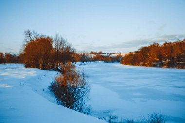 Gün batımında nehirde kış ormanı. Karlı ağaçlarla dolu renkli bir manzara, suda yansıması olan güzel donmuş bir nehir. Mevsimlik. Kış ağaçları, göl ve mavi gökyüzü. Buz gibi karlı nehir. Hava Durumu