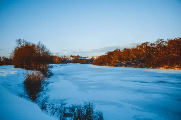Gün batımında nehirde kış ormanı. Karlı ağaçlarla dolu renkli bir manzara, suda yansıması olan güzel donmuş bir nehir. Mevsimlik. Kış ağaçları, göl ve mavi gökyüzü. Buz gibi karlı nehir. Hava Durumu