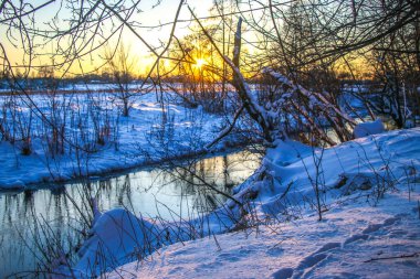 Karlı kıyıları olan bir nehre kar yağıyor. Suyun üzerinde ağaç dalları, güneşli bir günde ağaçlar karla kaplı. Ukrayna Shostka, Shostka nehri, Sunset.