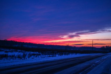 Ağaçlı ve tarla yollu kış günbatımı manzarası. Parlak kırmızı kış günbatımı, bulutların çizgileri, ağaçların siluetleri