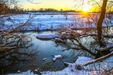 Karlı kıyıları olan bir nehre kar yağıyor. Suyun üzerinde ağaç dalları, güneşli bir günde ağaçlar karla kaplı. Ukrayna Shostka, Shostka nehri, Sunset.