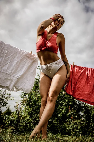 洗濯物を干した 若い美しいセクシーな女性は服を洗い 庭と晴れた日に屋外で下着を掛けます 赤と白のパンティーに身を包んだ — ストック写真