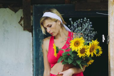 Kırmızı elbiseli güzel bir kadın eski ahşap bir evin yanındaki masada otururken elinde büyük bir buket ayçiçeğiyle bir demlik tutuyor.