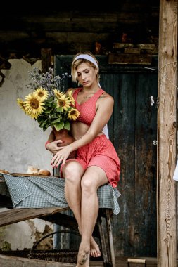 Kırmızı elbiseli güzel bir kadın eski ahşap bir evin yanındaki masada otururken elinde büyük bir buket ayçiçeğiyle bir demlik tutuyor.
