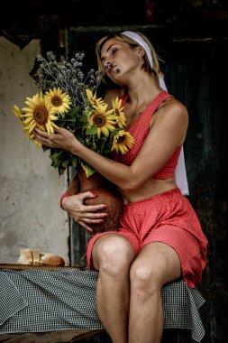 Kırmızı elbiseli güzel bir kadın eski ahşap bir evin yanındaki masada otururken elinde büyük bir buket ayçiçeğiyle bir demlik tutuyor.