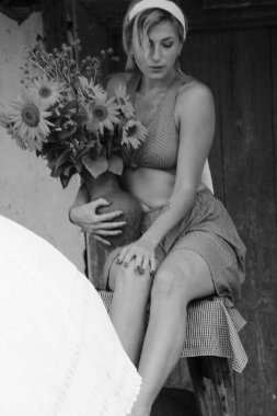 Kırmızı elbiseli güzel bir kadın, eski ahşap bir evin yanındaki masada otururken elinde büyük bir demet ayçiçeğiyle bir demlik tutuyor. Siyah beyaz bir fotoğraf.