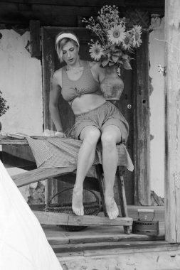 Kırmızı elbiseli güzel bir kadın, eski ahşap bir evin yanındaki masada otururken elinde büyük bir demet ayçiçeğiyle bir demlik tutuyor. Siyah beyaz bir fotoğraf.