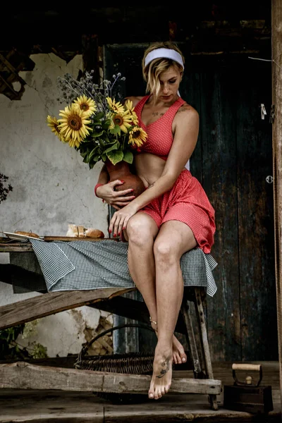 赤い服の美しい若い女性が古い木造の家の近くのテーブルの上に座っている間 ひまわりの大きな花束を持つ鍋を保持しています — ストック写真