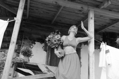 Kırmızı elbiseli güzel bir kadın, eski ahşap bir evin yanındaki masada otururken elinde büyük bir demet ayçiçeğiyle bir demlik tutuyor. Siyah beyaz bir fotoğraf.