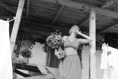 Kırmızı elbiseli güzel bir kadın, eski ahşap bir evin yanındaki masada otururken elinde büyük bir demet ayçiçeğiyle bir demlik tutuyor. Siyah beyaz bir fotoğraf.