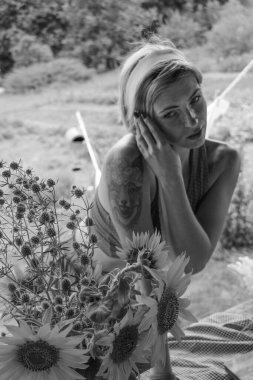 Kırmızı elbiseli güzel bir kadın, eski ahşap bir evin yanındaki masada otururken elinde büyük bir demet ayçiçeğiyle bir demlik tutuyor. Siyah beyaz bir fotoğraf.