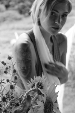 Kırmızı elbiseli güzel bir kadın, eski ahşap bir evin yanındaki masada otururken elinde büyük bir demet ayçiçeğiyle bir demlik tutuyor. Siyah beyaz bir fotoğraf.