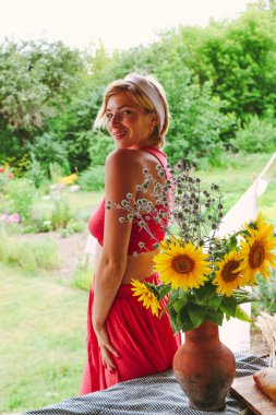 Kırmızı elbiseli güzel bir kadın portresi, büyük bir buket ayçiçeği ile bir demlik, eski ahşap bir evin yanında bir masada otururken
