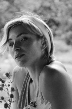 Kırmızı elbiseli güzel genç bir kadın, büyük bir buket ayçiçeği ile bir demlik tutuyor, eski ahşap bir evin yanında otururken siyah beyaz bir fotoğraf