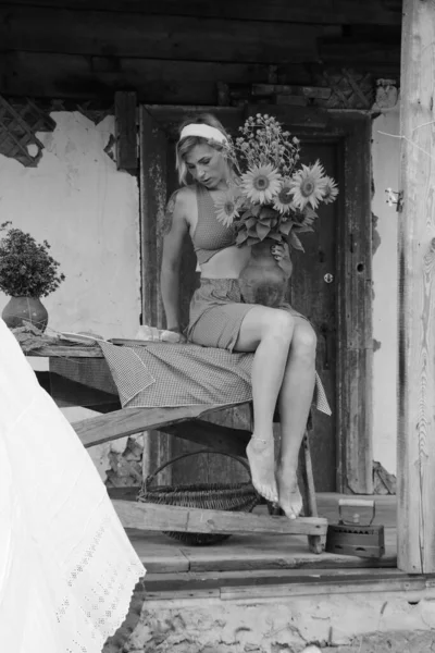 Kırmızı elbiseli güzel bir kadın, eski ahşap bir evin yanındaki masada otururken elinde büyük bir demet ayçiçeğiyle bir demlik tutuyor. Siyah beyaz bir fotoğraf.