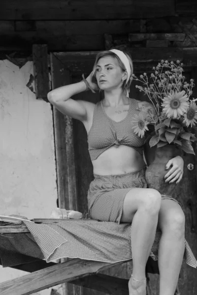 赤い服の美しい若い女性は 古い木造の家の近くのテーブルの上に座っている間 ひまわりの大きな花束を持つ鍋を保持黒と白の写真 — ストック写真