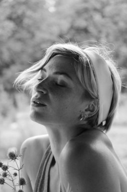 Kırmızı elbiseli güzel genç bir kadın, büyük bir buket ayçiçeği ile bir demlik tutuyor, eski ahşap bir evin yanında otururken siyah beyaz bir fotoğraf