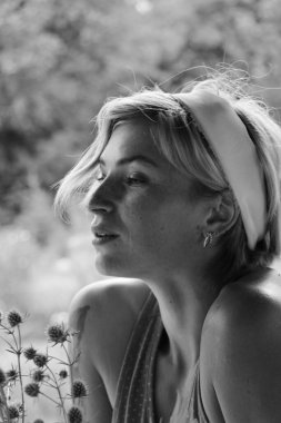 Kırmızı elbiseli güzel genç bir kadın, büyük bir buket ayçiçeği ile bir demlik tutuyor, eski ahşap bir evin yanında otururken siyah beyaz bir fotoğraf