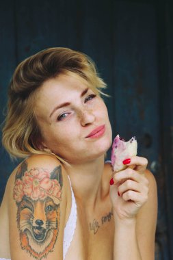 Sarışın, komik, seksi bir kadının portresi. Eski bir köy evinin verandasındaki masada yemek yiyor. Bir buket ayçiçeği..