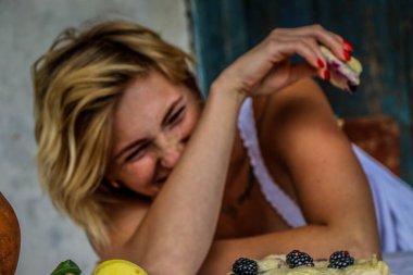 Sarışın, komik, seksi bir kadının portresi. Eski bir köy evinin verandasındaki masada yemek yiyor. Bir buket ayçiçeği..