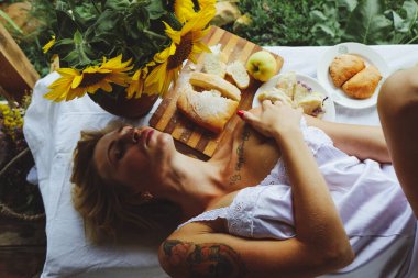 Güzel, genç, seksi, komik bir kızın portresi. Mutfak masasında yatan sarışın. Masada yemek, meyve ve çiçeklerin yanında yatıyor, yiyor.