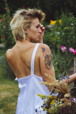 Beyaz elbiseli doğal sarışın seksi Avrupalı kız bahçe çiçekleriyle dolu bir sepet tutuyor. Bir yaz günü bahçede yürür