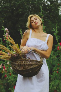 Beyaz elbiseli doğal sarışın seksi Avrupalı kız bahçe çiçekleriyle dolu bir sepet tutuyor. Bir yaz günü bahçede yürür