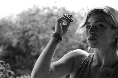 Kırmızı elbiseli güzel bir kadın, eski ahşap bir evin yanındaki masada otururken elinde büyük bir buket ayçiçeği olan bir çömlek tutuyor, siyah beyaz bir fotoğraf.