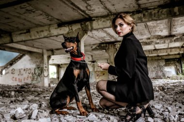 Çekici, genç, çekici, baştan çıkarıcı, siyah deri elbiseli bir kadın. Eski bir binanın yıkıntıları üzerinde. Yanında güzel bir köpek, Doberman..
