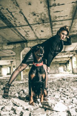 Çekici, genç, çekici, baştan çıkarıcı, siyah deri elbiseli bir kadın. Eski bir binanın yıkıntıları üzerinde. Yanında güzel bir köpek, Doberman..