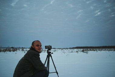 Telefonlu bir adam Zidio 'nun kış günbatımının hızlandırılmış fotoğraflarını çekiyor. Tarla, yol ve ağaçlar karla kaplı..