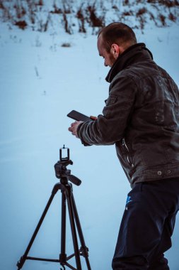 Telefonlu bir adam Zidio 'nun kış günbatımının hızlandırılmış fotoğraflarını çekiyor. Tarla, yol ve ağaçlar karla kaplı..