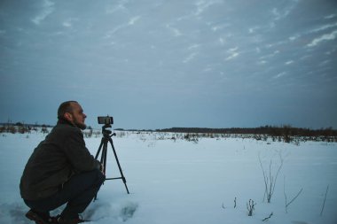 Telefonlu bir adam Zidio 'nun kış günbatımının hızlandırılmış fotoğraflarını çekiyor. Tarla, yol ve ağaçlar karla kaplı..