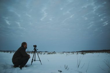 Telefonlu bir adam Zidio 'nun kış günbatımının hızlandırılmış fotoğraflarını çekiyor. Tarla, yol ve ağaçlar karla kaplı..
