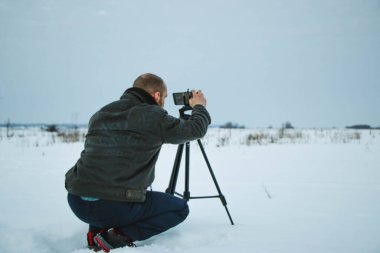 Telefonlu bir adam Zidio 'nun kış günbatımının hızlandırılmış fotoğraflarını çekiyor. Tarla, yol ve ağaçlar karla kaplı..