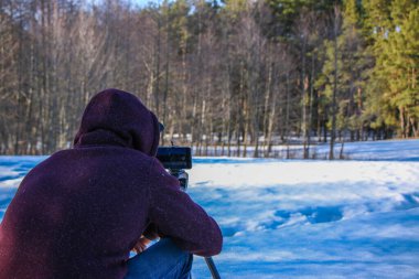 Bir blog için içerik oluşturulurken modern özgürlük iş başında. Erkek turist gezgin blogcu kışlık kıyafetlerle telefonda video çekiyor. Karlı, güneşli bir günün doğal bir bölgesinde duruyor..