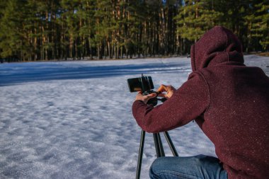 Bir blog için içerik oluşturulurken modern özgürlük iş başında. Erkek turist gezgin blogcu kışlık kıyafetlerle telefonda video çekiyor. Karlı, güneşli bir günün doğal bir bölgesinde duruyor..