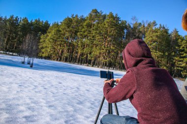 Bir blog için içerik oluşturulurken modern özgürlük iş başında. Erkek turist gezgin blogcu kışlık kıyafetlerle telefonda video çekiyor. Karlı, güneşli bir günün doğal bir bölgesinde duruyor..