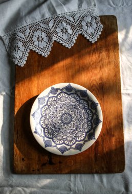 Beyaz keten bir masa örtüsü üzerinde eski ahşap bir tahta ve mavi desenli dantel havluyla beyaz bir tabak. Güneşli doğal ışık, gölge. Mutfak masası manzaralı. Güzel retro, rustik arkaplan, serbest alan.