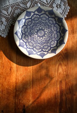 Beyaz keten bir masa örtüsü üzerinde eski ahşap bir tahta ve mavi desenli dantel havluyla beyaz bir tabak. Güneşli doğal ışık, gölge. Mutfak masası manzaralı. Güzel retro, rustik arkaplan, serbest alan.