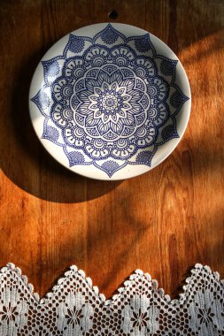 Beyaz keten bir masa örtüsü üzerinde eski ahşap bir tahta ve mavi desenli dantel havluyla beyaz bir tabak. Güneşli doğal ışık, gölge. Mutfak masası manzaralı. Güzel retro, rustik arkaplan, serbest alan.