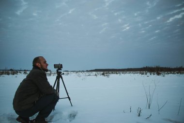 Telefonlu bir adam Zidio 'nun kış günbatımının hızlandırılmış fotoğraflarını çekiyor. Tarla, yol ve ağaçlar karla kaplı..